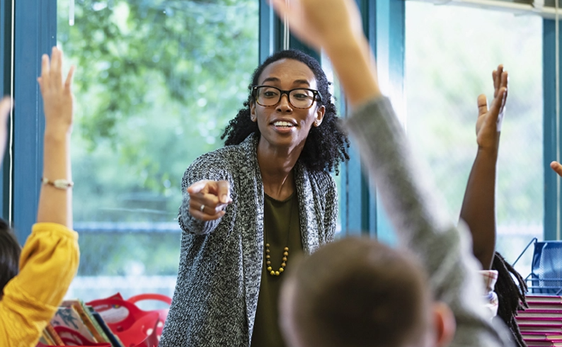 A teacher pointing to one of her students who is raising their hand in class.