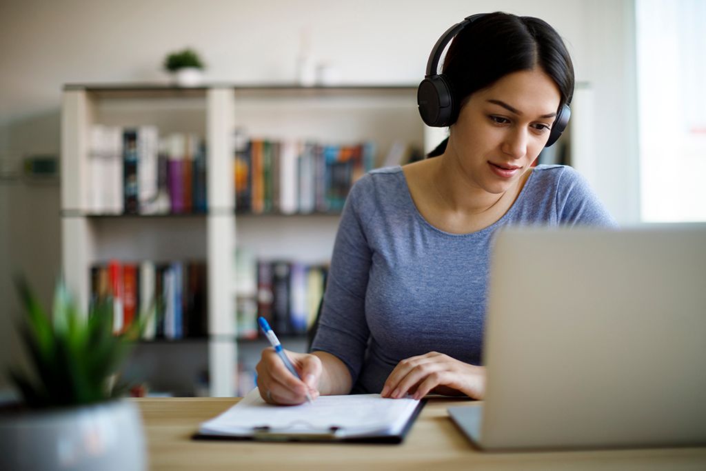 A woman is studying at a desk. She is writing notes while looking at a laptop.