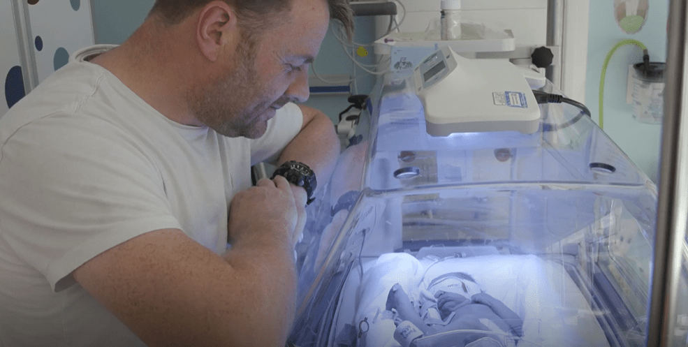 A father looking at his infant child inside of an incubator.
